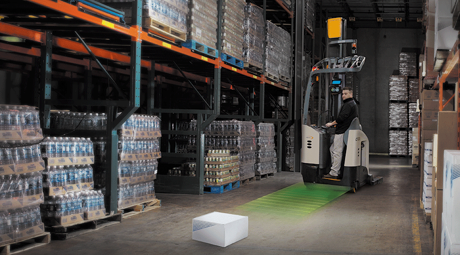 A worker drives a forklift toward a white box on the floor in a warehouse.
